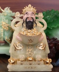 Thần Tiền ngồi trên những thỏi vàng lấp lánh, tay ôm thỏi vàng lớn tượng trưng cho giàu sang phú quý
